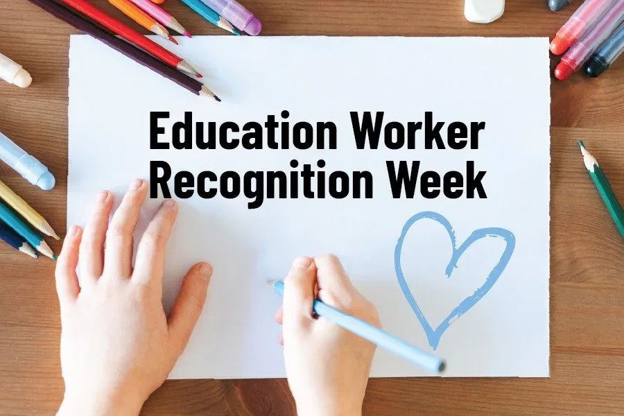 Child's hands writing on a canvas that states Education Worker Recognition Week. Child's hands are surrounded by pencil crayons with everything laying on a desk