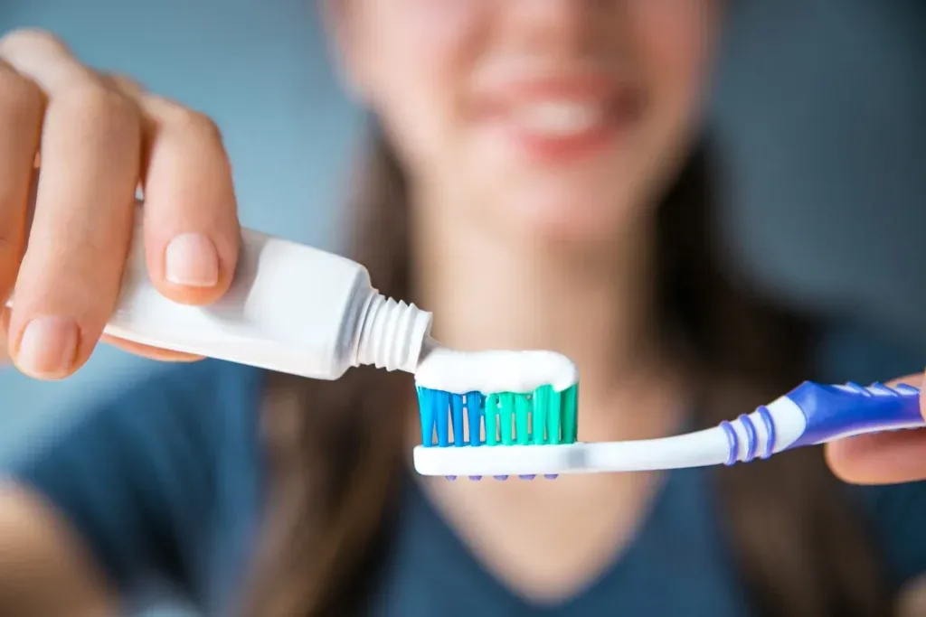Photo focuses on a blue and white tooth brush and toothpaste as a woman in the background smiles and puts the toothpaste on the brush