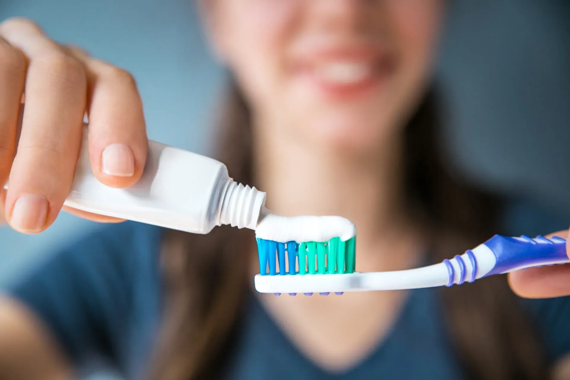 Photo focuses on a blue and white tooth brush and toothpaste as a woman in the background smiles and puts the toothpaste on the brush